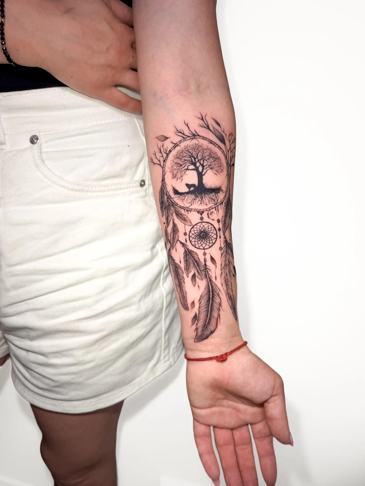 black & grey dreamcatcher with a tree and small wolf silhouette inside the hoop, hanging feathers and a small web, on inner forearm