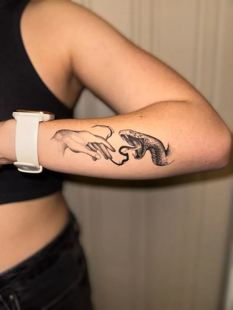 black & grey shaded hand reaching toward an open-mouthed snake with a forked tongue, on the outer forearm below the elbow