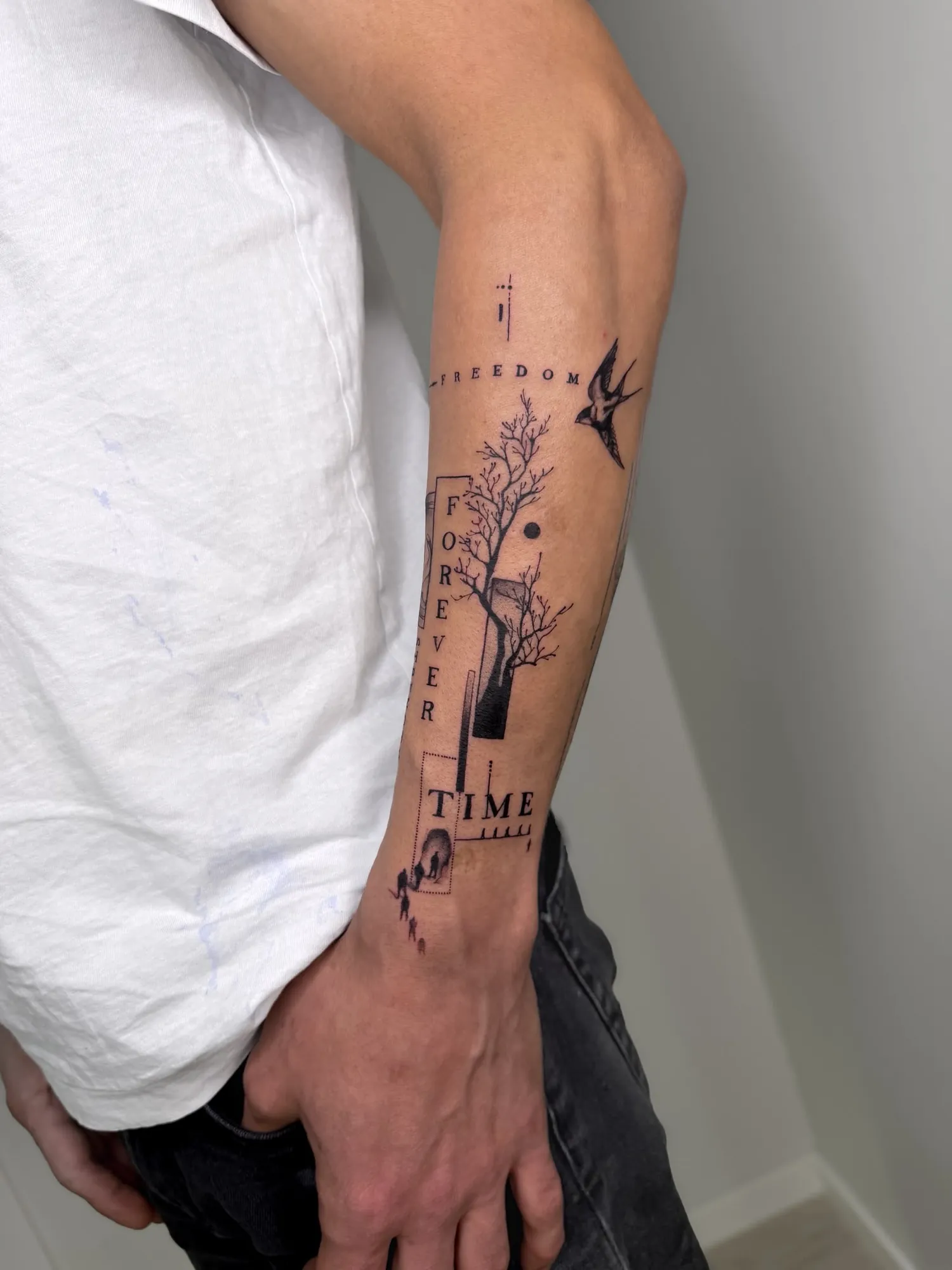 blackwork forearm collage with a bare tree, a swallow, small human silhouettes and an elephant, and the words 'FREEDOM', 'FOREVER' (vertical) and 'TIME' in serif capitals, on the outer forearm