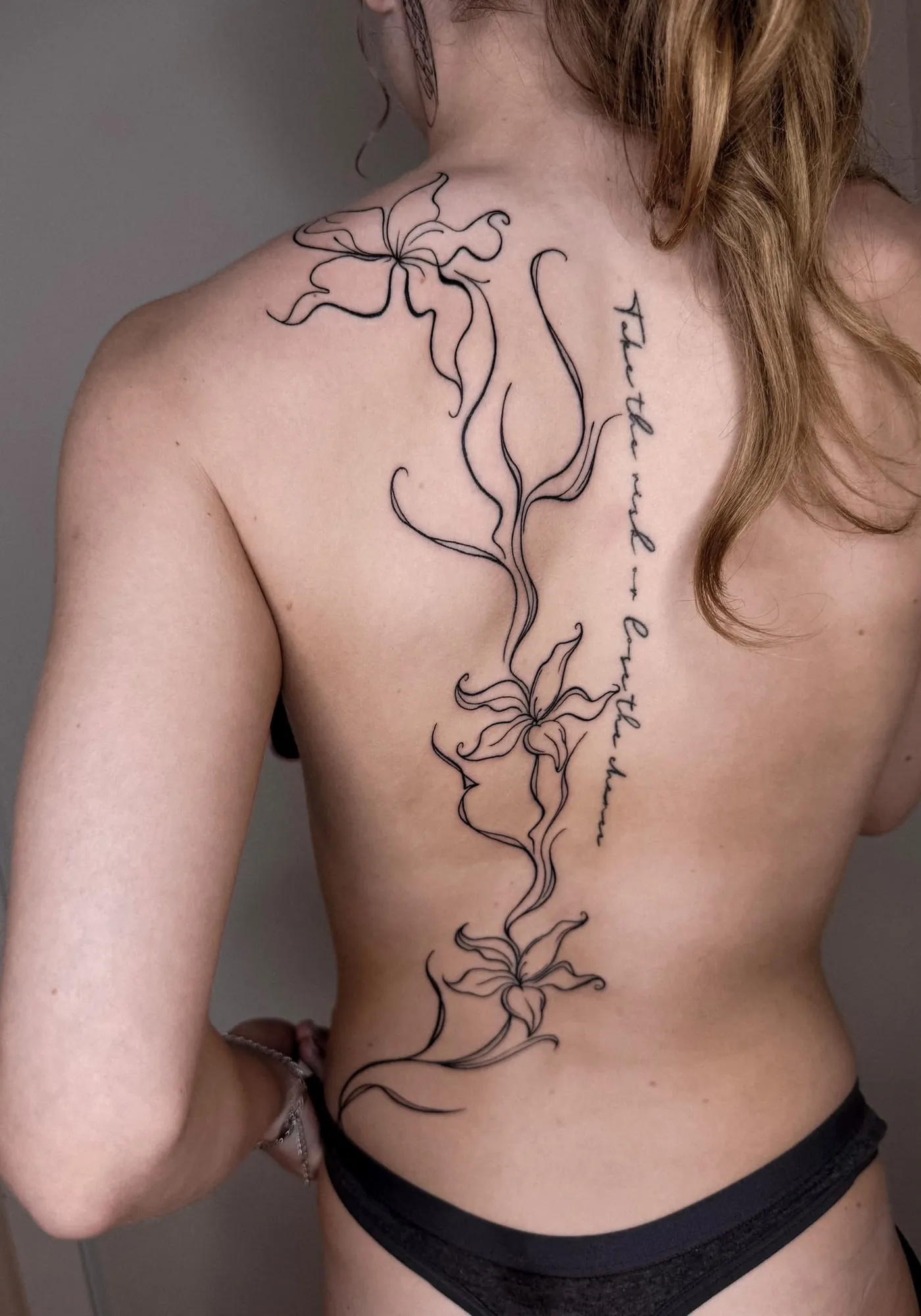 large fineline lily-type flowers running from the shoulder blade down the spine to the lower back, with cursive script beside the spine reading 'Take the risk or lose the chance' (small, partly hard to read)