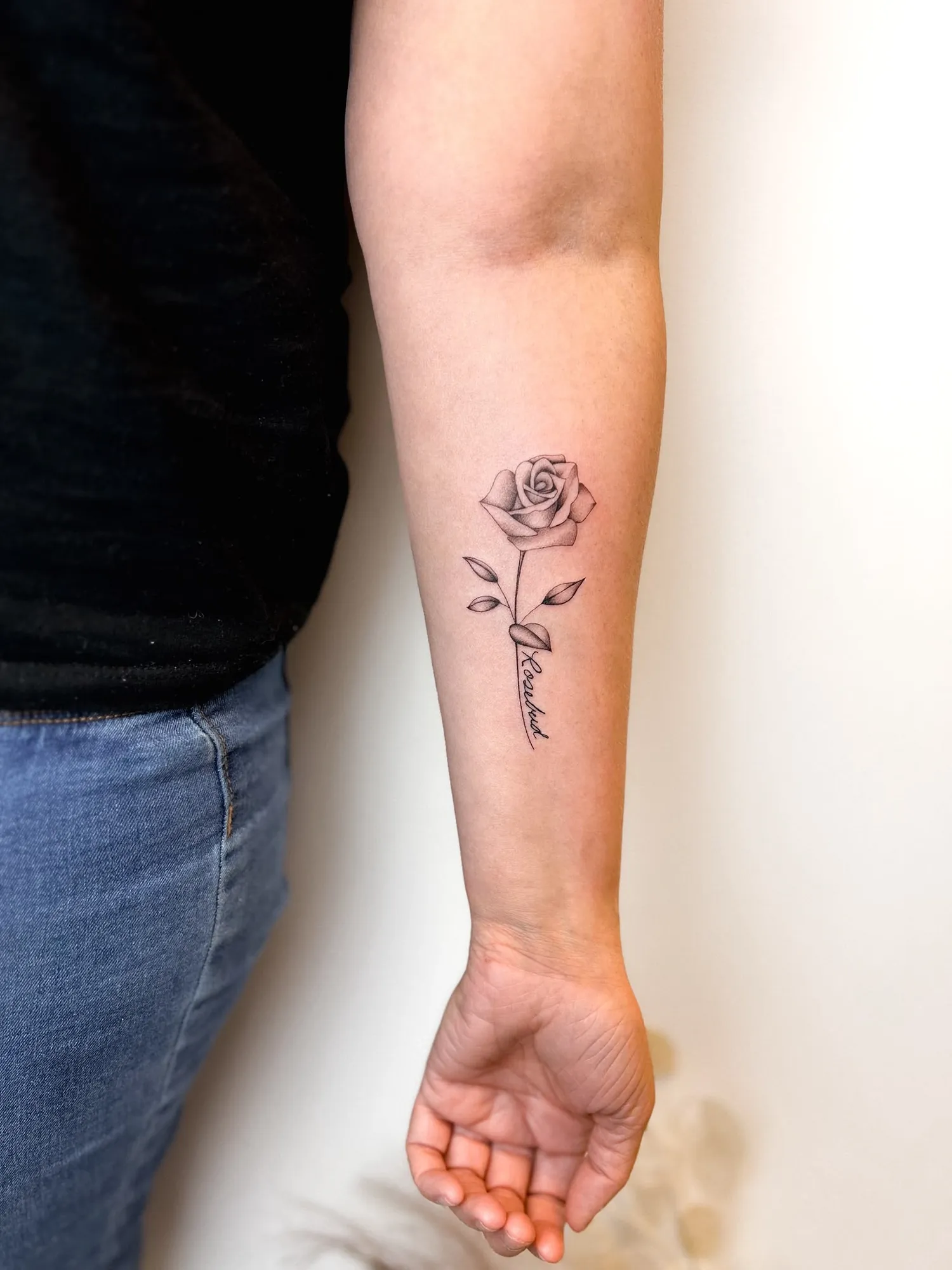 black & grey fineline rose with leaves and stem, script 'Rosebud' written along the stem, on inner forearm