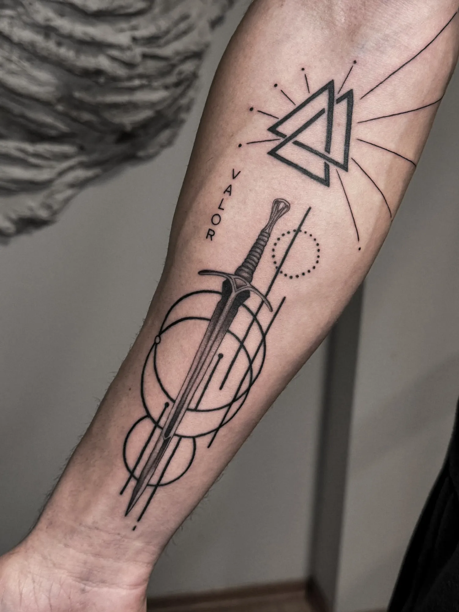 black & grey shaded sword with bold geometric circles and lines, a valknut of three interlocking triangles with rays, and the word 'VALOR' on inner forearm