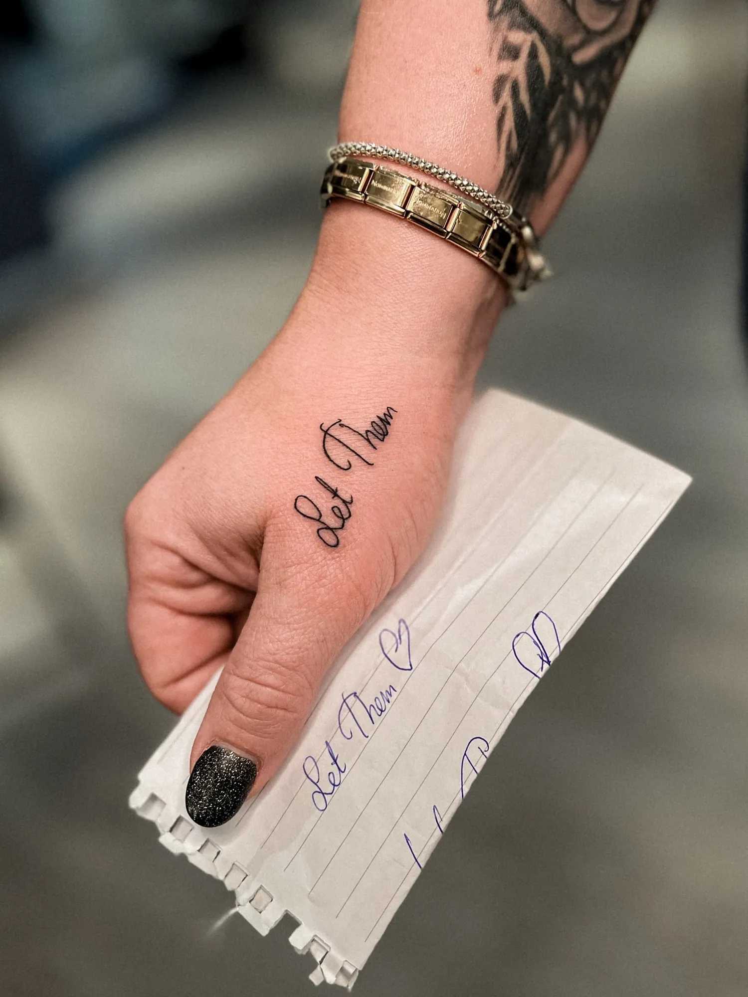 cursive script 'Let Them' on the side of the hand below the index finger, shown next to the handwritten paper reference